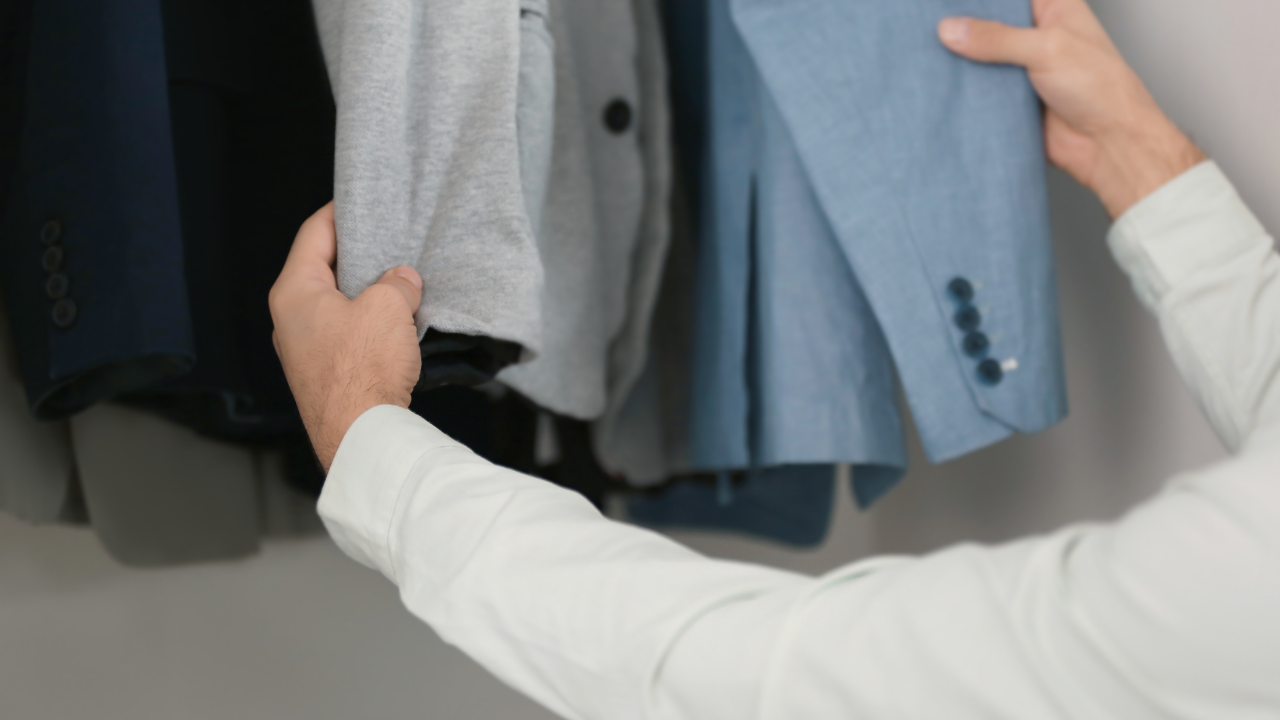 A person wearing a white shirt is selecting between a grey shirt and a light blue blazer from a row of hanging clothes in a closet, guided by the expertise of a men's wardrobe stylist.